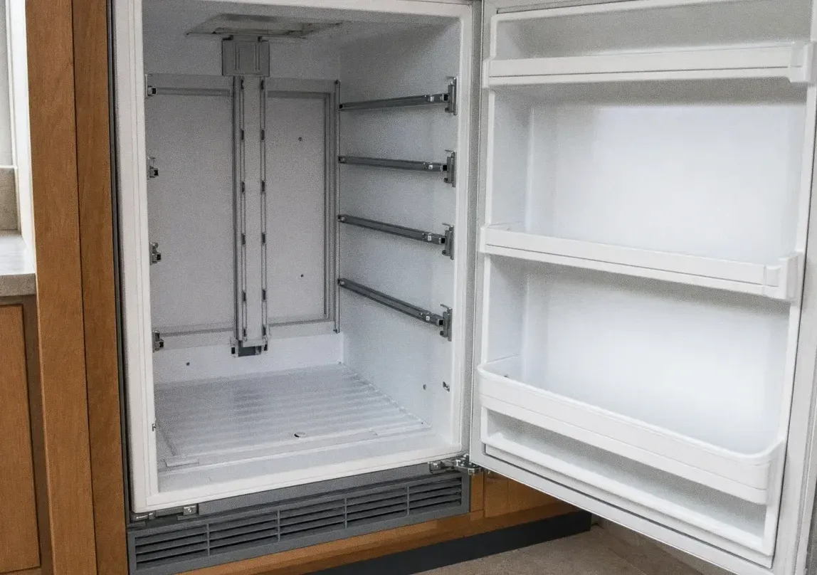 An under-counter refrigerator with its door open on an empty white interior and bare shelf support rails